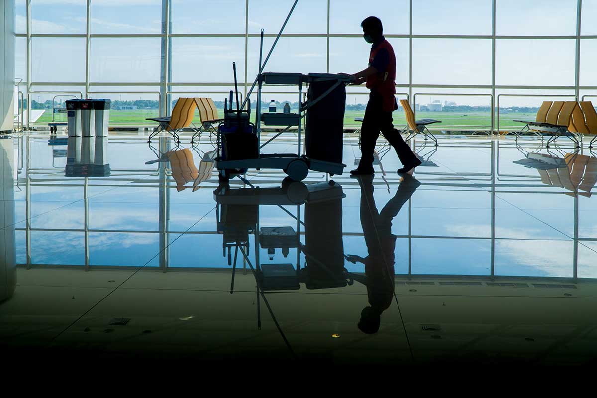 Man walking through an airport while pushing a cleaning equipment cart with large windows in the background.