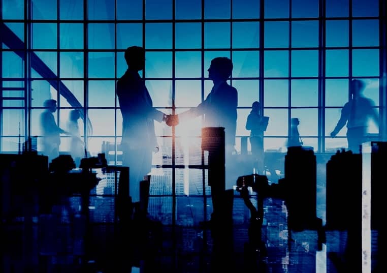 Silhouettes of two businessmen shaking hands in a glass office building as people walk in the background